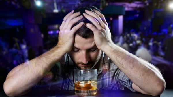 young alcoholic drunk man depressed, sad, wasted and low thinking about alcohol addiction problem and drinking indoors at bar of a disco nightclub hands on his head in front of whiskey glass in alcoholism concept